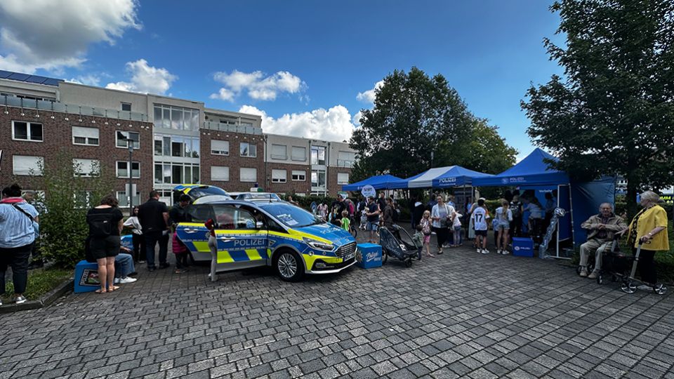 Einsatzfahrzeug im Vordergrund, Gebäude und Personen, blauer Himmel mit Schönwetterwolken