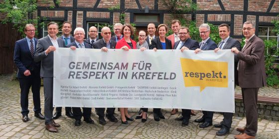 Die Leiterinnen und Leiter aller mitwirkenden Institutionen halten ein Banner mit der Aufschrift: Gemeinsam für Respekt in Krefeld. Ein Fachwerkhaus (Zooscheune) befindet sich im Hintergrund.